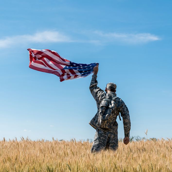 A male American Soldier standing in a field holding the American flag up waving in the wind their their back turned.